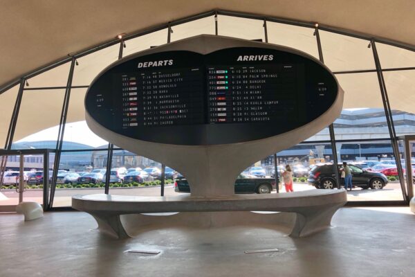 A curved airport departures and arrivals board above a concrete bench in a bright terminal